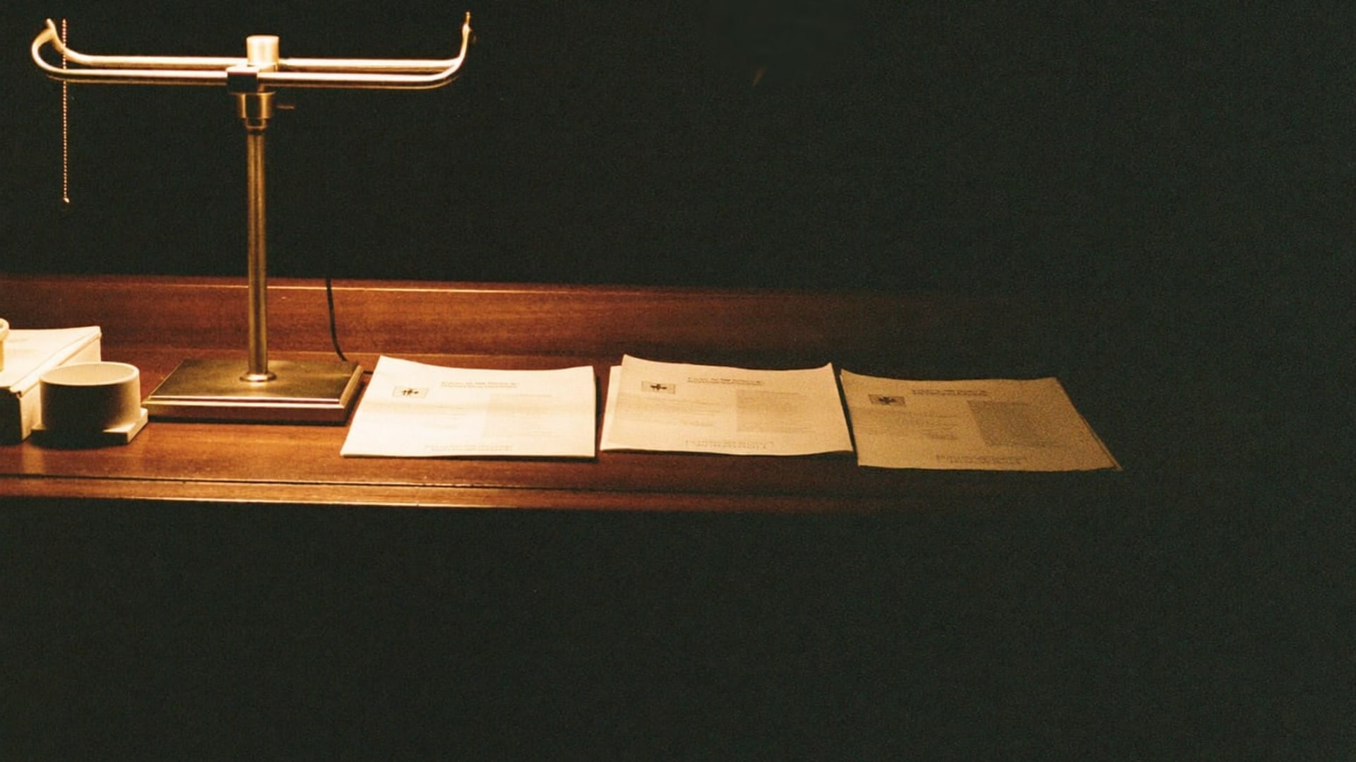 The same front desk before dawn, framed wide — the lamp, the printed lists, the dark room waiting for the first guest.
