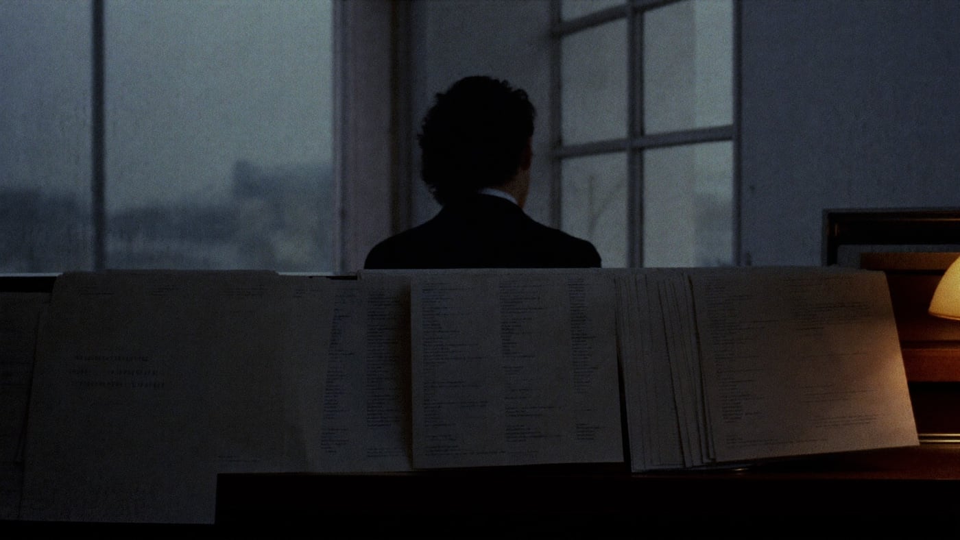 A receptionist in silhouette at a cold pre-dawn window, seen from behind over thick stacks of printed guest lists — the paper morning.